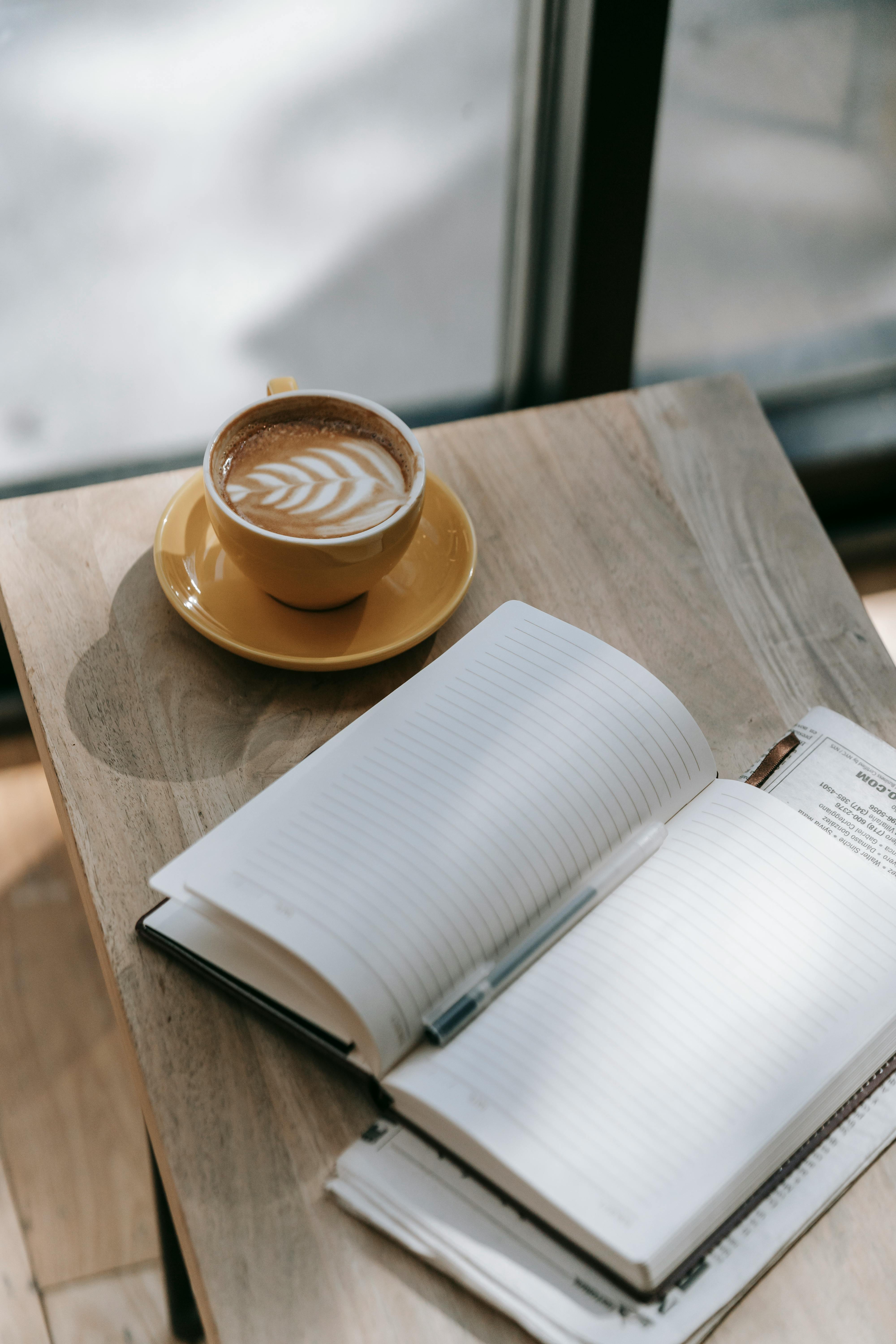 Kop koffie met latte art op een houten tafel naast een open notitieboekje